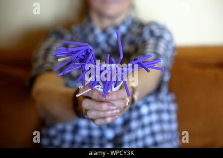 Plastic violet drinking straws, disposable, holds in senior woman hands, close up, person in soft focus sit on couch. Stock Photo