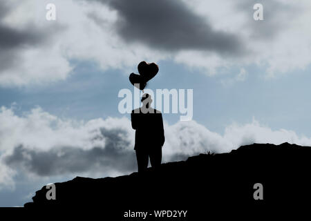 A Man with heart balloon. Symbols. A man with two hearts balloon stands on a hill in front of blue sky with clouds Stock Photo