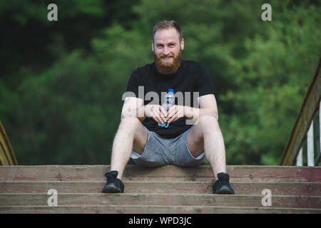 happy smiling bearded man with bottle of water sitting on wooden bridge in autumn green outdoor park Stock Photo