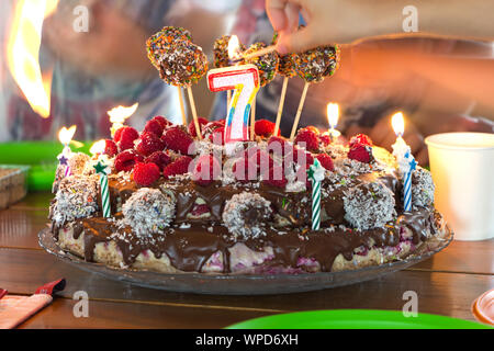 Setting fire to a candles on a childrens birthday cake. Selective focus on holiday candle in form of number 7, hands with burning matches in bokeh. Un Stock Photo