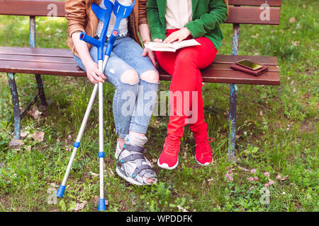 Two friends reading a book in the park, one has a broken foot Stock Photo