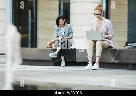 Full length portrait of two young businesswoman using laptops while working outdoors in urban setting, copy space Stock Photo