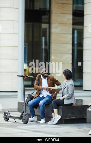 Portrait of smiling African-American man shaking hands with Asian woman while sitting on bench outdoors, copy space Stock Photo