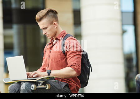 Side view portrait of handsome young man using laptop while studying outdoors in urban setting, copy space Stock Photo