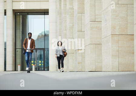 Full length portrai of two people walking in city street, focus on African-American man riding electric scooter, copy space Stock Photo