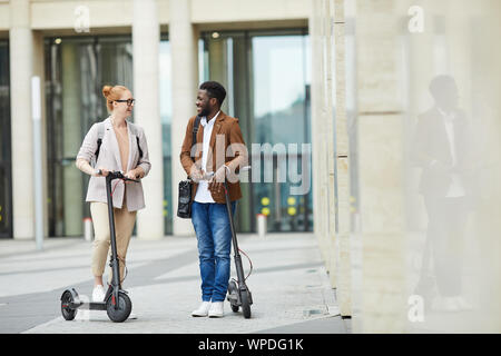 Full length portrait of mixed race young couple riding alectric scooters in city street and smiling happily, copy space Stock Photo