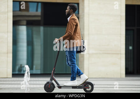 Side view full length of modern African-American businessman riding electric scooter while commuting to work in city, copy space