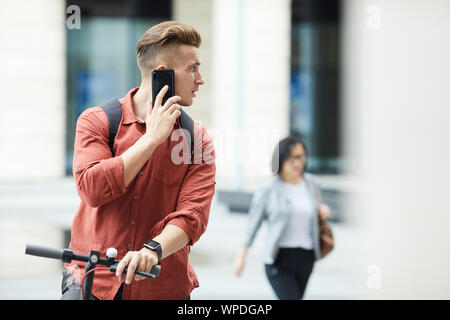 Waist up portrait of handsome young man speaking by smartphone while riding electric scooter in urban setting, copy space