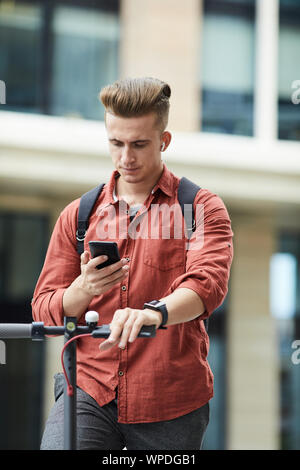 Waist up portrait of handsome young man checking smartphone while riding electric scooter in urban setting, copy space Stock Photo