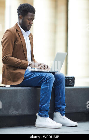 Full length side view of young African-American man using laptop outdoors while working on freelance project in urban setting, copy space Stock Photo
