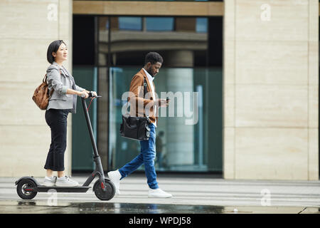 Full length portrait of people commuting in city, focus on Asian woman riding electric scooter in foreground, copy space Stock Photo