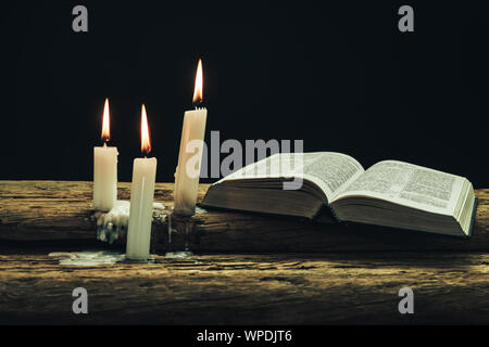 Beautiful open bible and burned candles on a old oak wooden table and dark background. Stock Photo