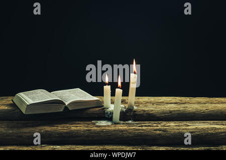 Beautiful open bible and burned candles on a old oak wooden table and dark background. Stock Photo