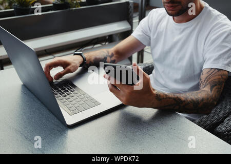Handsome bearded guy with tattooed hands dressed white shirt using smartphone and notebook computer in outdoors coffee shop, male holding cell phone a Stock Photo