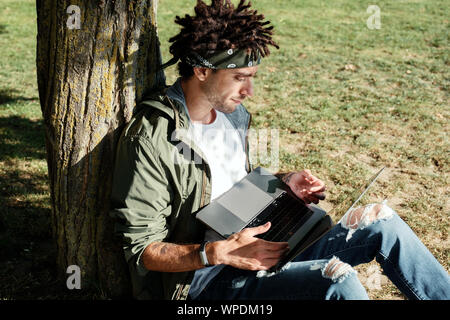 Young millennial male student with dreadlocks using laptop outdoor in public park, programmer working remotely on notebook, searching information usin Stock Photo