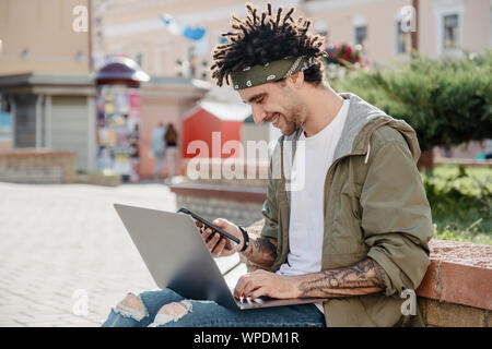 Side view shot of a handsome man using smartphone while working on laptop outdoor, happy guy with african dreadlocks hairstyle check notification, rea Stock Photo