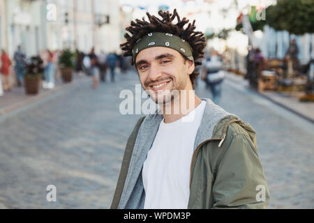 Young handsome beard man dressed stylish casual clothes, white t-shirt, green jacket, bandana posing outdoor. Smiling hipster with dreadlocks hairstyl Stock Photo