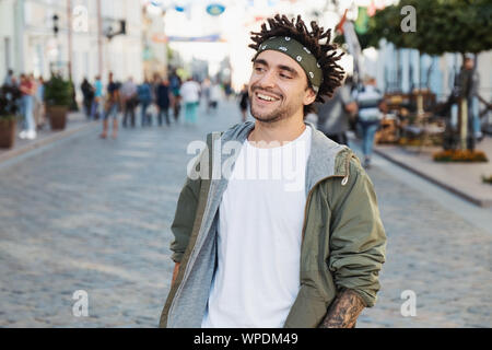 Young handsome beard man dressed stylish casual clothes, white t-shirt, green jacket, bandana posing outdoor. Smiling hipster with dreadlocks hairstyl Stock Photo