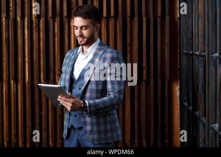 young successful businessman with a beard in a luxurious blue checked suit. Man works on a tablet in a loft office. Stock Photo