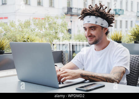 Cheerful bearded hipster guy with dreadlocks hairstyle working on laptop in outdoor street cafe. Freelancer, it professional in coworking space, remot Stock Photo