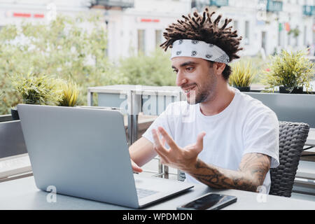 Happy bearded hipster guy waving hand and talking on video chat using wireless network on laptop device. Handsome man with dreadlocks hairstyle vloggi Stock Photo