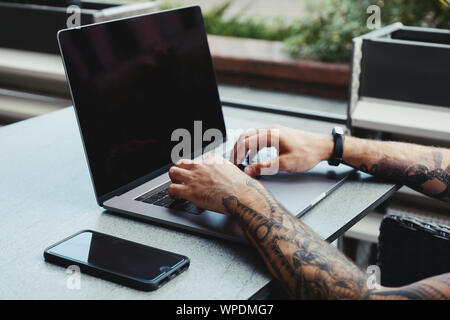 Young tattooed man working on laptop in a coffee shop. Rear view of hipster hands with tattoo busy using laptop at coworking office desk. Guy typing t Stock Photo