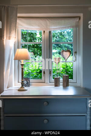 Window inside country house with lamp and heart decoration on chest of drawers. Stock Photo