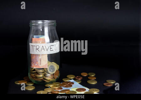 Glass transparent jar labeled handwritten travel. Euro Banknotes and coins inside and outside of the jar. Isolated black background. Stock Photo