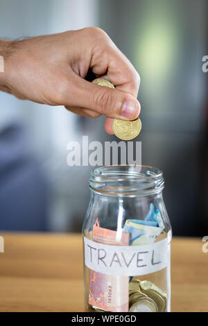 Man's hand holding a coin to put on a jar handwritten labeled travel to save for a trip. Stock Photo