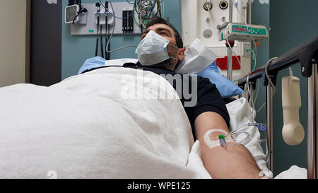 Young man lying in hospital bed. Recovering in modern hospital wards, covered with blanket, face mask and with intravenous needle in his arm