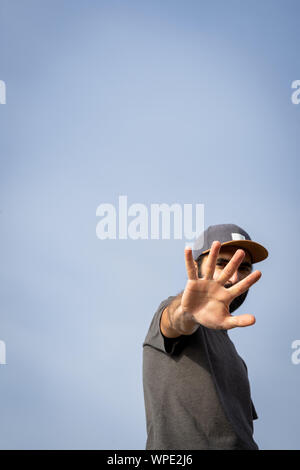 Bearded Man wearing a cap, view from the front, with the arm stretched to the camera with the palm of his hand opened covering his face partially. Stock Photo