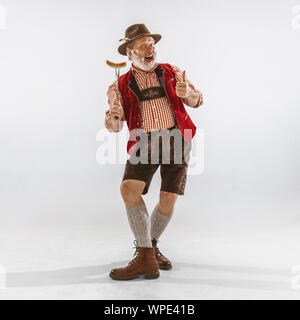 Portrait of Oktoberfest senior man in hat, wearing the traditional Bavarian clothes. Male full-length shot at studio on white background. The celebration, holidays, festival concept. Eating sausage. Stock Photo
