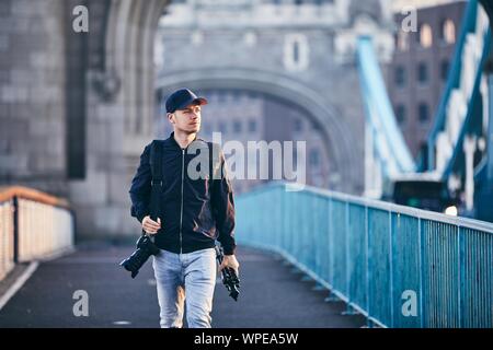 Photographer looking away while carrying camera and tripod against Tower Bridge. London, United Kingdom. Stock Photo