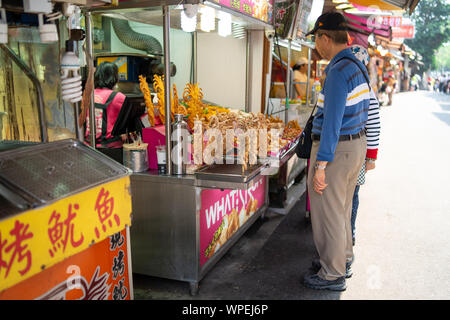 Elderly couple buying traditional crispy deep fried squid at a street food stall on the Danshui (Tamsui) main street at day. Stock Photo