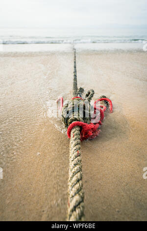 Creative red knotted ships rope lying on a sandy beach leading out to sea on the shoreline as the tide comes in. Stock Photo