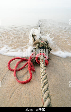 Creative red knotted ships rope lying on a sandy beach leading out to sea on the shoreline as the tide comes in. Stock Photo