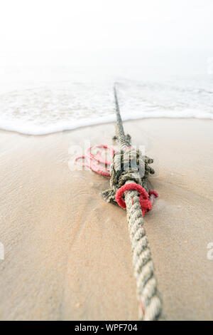 Creative red knotted ships rope lying on a sandy beach leading out to sea on the shoreline as the tide comes in. Stock Photo