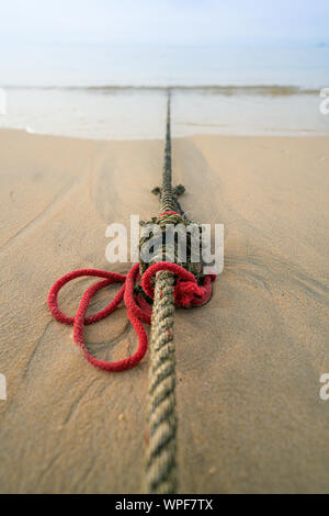 Creative red knotted ships rope lying on a sandy beach leading out to sea on the shoreline as the tide comes in. Stock Photo