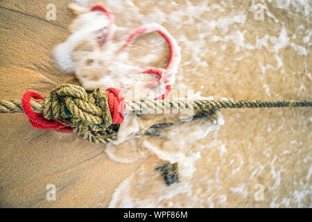 Creative red knotted ships rope lying on a sandy beach leading out to sea on the shoreline as the tide comes in. Stock Photo