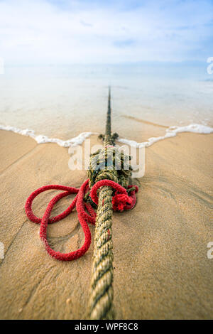 Creative red knotted ships rope lying on a sandy beach leading out to sea on the shoreline as the tide comes in. Stock Photo