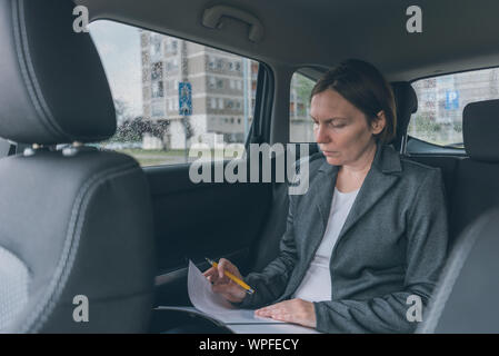 Businesswoman doing business paperwork on car back seat, adult caucasian female businessperson executive analyzing business results while traveling by Stock Photo