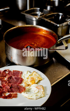closeup of a white ceramic plate with some slices of serrano ham and some fried eggs next to a saucepan with tomato sauce, on the counter of a profess Stock Photo