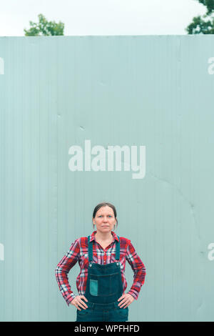 Serious female farmer posing on farm. Confident woman farm worker wearing plaid shirt and jeans overalls looking at camera, with copy space Stock Photo