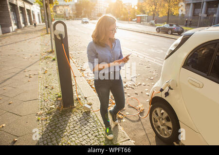 Young woman is standing near the electric car and looks at the smart phone. The rental car is charging at the charging station for electric vehicles. Stock Photo