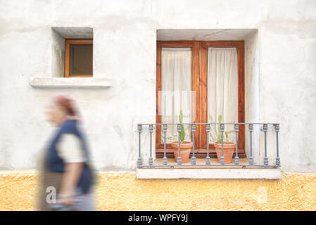 Old woman walking down the street in Andalusia, Spain against a balcony decorated with flowerpots and geraniums. Empty copy space for Editor's text. Stock Photo