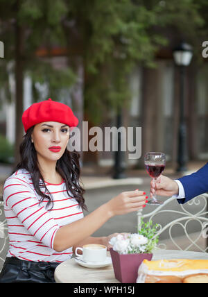 Elegant man in a suit gives a pretty brunette woman in a red cap and striped t-shirt glass of red wine in outdoor cafe. Stock Photo