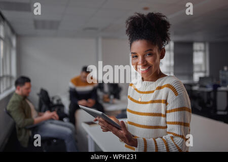 Cheerful young african american businesswoman in casual wear holding digital tablet smiling and looking to camera while her colleagues communicating Stock Photo