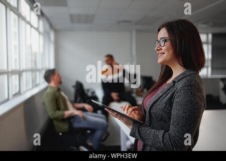 Thoughtful young businesswoman looking away while holding digital tablet in front of colleagues relaxing in office at background Stock Photo