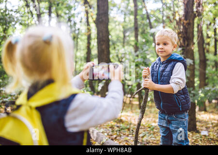 Children preschoolers Caucasian brother and sister take pictures of each other on mobile phone camera in forest park autumn. theme of hobby and active Stock Photo