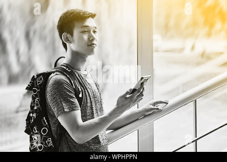 Young asian male student with smartphone in hand wearing a t-shirt and a backpack standing on university stairs and looks optimistic into future in Stock Photo